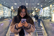 © Julio Rodriguez/Westend61 - Young woman using smart phone while standing against illuminated glass wall in shopping mall