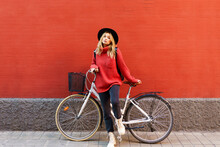 Young Blond Woman Wearing Hat Standing With Bicycle Against Red Wall