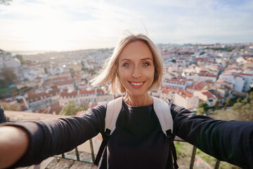 Wall Mural - Traveling by Portugal. Young traveling woman taking selfie in old town Lisbon with view on red tiled roofs, ancient architecture.