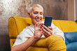 © OneInchPunch/Westend61 - Cheerful senior woman using smart phone while sitting on sofa in living room