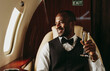 © OneInchPunch/Westend61 - Smiling male entrepreneur holding champagne looking out of window in airplane