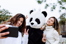 Cheerful Female Friends Taking Selfie With Male Friend In Panda Mask While Gesturing Peace Sign At Park