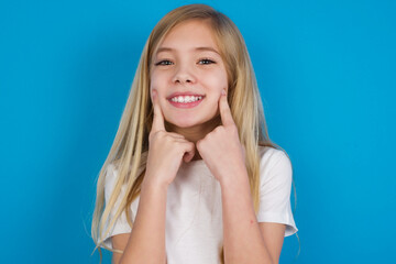 Wall Mural - Happy beautiful Caucasian little girl wearing white T-shirt over blue background with toothy smile, keeps index fingers near mouth, fingers pointing and forcing cheerful smile