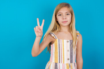 Wall Mural - beautiful Caucasian little girl wearing stripped dress over blue background showing and pointing up with fingers number two while smiling confident and happy.