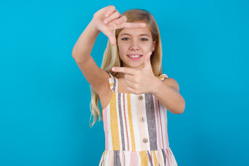 Wall Mural - Positive beautiful Caucasian little girl wearing stripped dress over blue background with cheerful expression, has good mood, gestures finger frame actively at camera.