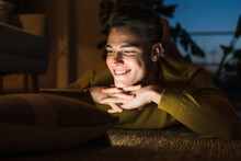 Smiling Young Man Lying On Carpet While Watching Videos Through Digital Tablet In Living Room During Night
