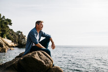 Thoughtful Mature Man Sitting On Rock Against Clear Sky