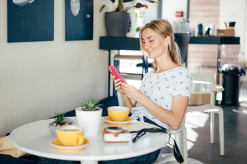 Wall Mural - Portrait of a young beautiful caucasian stylish blonde sitting at a table in a coffee shop. Woman laughing while chatting in the mobile phone.