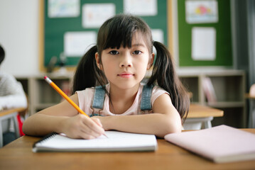 Wall Mural - Portrait of little Asian girl writing or drawing in notebook at desk in classroom and looking at camera at the elementary school. Education knowledge, activities with kids concept.