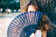 Horizontal portrait of a brunette woman holding a blue hand fan in front of her face.