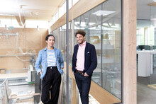 Male And Female Entrepreneurs With Hands In Pockets Standing By Glass Wall In Factory