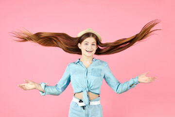 Portrait of cute pretty teenager wearing denim clothing and straw hat. Girl playing with long hair on pink background. Flying hairstyle.