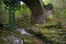 Old Roman Stone Bridge In The Forest In Tuscany. A Stream Flows Beneath It.