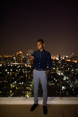 Wall Mural - Full length shot of African businessman outdoors at rooftop during night using phone