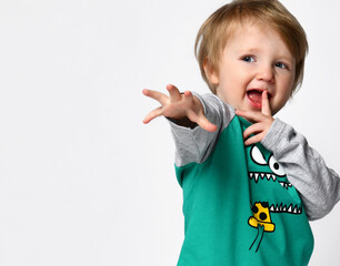 Little smiling preschool toddler boy European ethnicity wearing trendy green raglan with cute monster muzzle print and sportive black pants stand with hand reaching to camera. Studio portrait on white