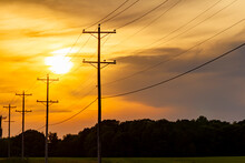 Silhouettes Of Utility Poles Carrying High Voltage Electricity Alongside A Road At A Rural Area. Sunset, Sunrise Image With Cloudy Orange Red Sky And Depth Of Field With Nobody In The Frame.