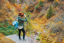 Female Tourist With A Backpack Admires The Mountain Scenery. A Red-haired Woman Ties Her Hair On The Edge Of A Cliff. Colosful Mountain Forest.