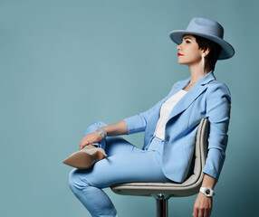 Wall Mural - Smiling brunette woman in blue business suit, hat and high-heeled shoes sits relaxed on chair looking at copy space over blue background. . Side view. Stylish female wear and fashion concept