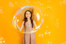 Little Happy Girl Playing With Huge Soap Bubbles On A Colored Yellow Background. The Child Have Fun Inside The Bubbles.