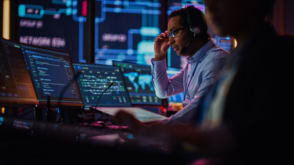 Professional IT Technical Support Specialist and Software Developer Working on Computer in Monitoring Control Room with Digital Screens. Employee Uses Headphones with Mic, Talking on a Call.