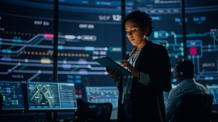 Fototapete - Young Multiethnic Female Government Employee Uses Tablet Computer in System Control Monitoring Center. In the Background Her Coworkers at Their Workspaces with Many Displays Showing Technical Data.