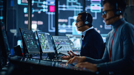 Wall Mural - Portrait of Professional IT Technical Support Specialist Working on Computer in Monitoring Control Room with Digital Screens. Employee Wears Headphones with Mic and Talking on a Call.