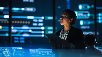 Fototapete - Young Multiethnic Female Government Employee Uses Tablet Computer in System Control Monitoring Center. In the Background Her Coworkers at Their Workspaces with Many Displays Showing Technical Data.