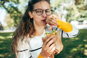 Wall Mural - Portrait of a student female has a delicious lunch with a sandwich outdoors. A blonde young woman in eyeglasses takes a rest to eat fast food in the park.