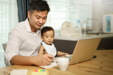 Work from home. Asian young father write notebook and holding his son on knee. looking at notebook. Entrepreneur man working with laptop. A boy sitting with dad at home in kitchen. quarantine time