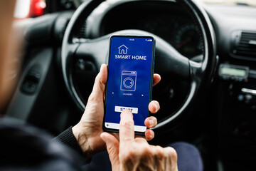 Crop anonymous person sitting on driver seat in car and using mobile phone with smart home app on screen while programming the washing machine remotely - - Internet of things (IOT) concept
