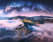 Milky Way arch and mountains in low clouds at starry night in summer. Landscape with pink sky with stars, arched Milky Way, trees on the hill in fog, mountain peaks. Space and galaxy. Aerial view 