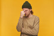 Stressed man, tired guy with blond hair, beard and mustache. Wearing green beanie and beige sweater. Touching bridge of nose in pain. Concentrating. Stand isolated over yellow background