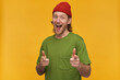 Portrait of positive, cheerful male with blond hairstyle and beard. Wearing green t-shirt and red beanie. Has tattoo. Pointing fingers at you. Watching at the camera isolated over yellow background