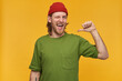 Portrait of handsome, happy male with blond hairstyle and beard. Wearing green t-shirt and red beanie. Has tattoo. Pointing thumb at himself. Winks at the camera isolated over yellow background