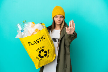 Wall Mural - Little girl holding a bag full of plastic bottles to recycle over isolated blue background making stop gesture