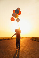 Wall Mural - Little girl holding colorful balloons in hand goes into the sunset