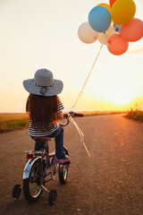 Wall Mural - Cute little girl with colorful balloons cycling at the road at sunset sky summer time. Back view.