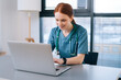 © dikushin - Cheerful smiling young female doctor in blue green medical uniform typing on laptop computer looking on screen sitting at desk near window in modern office of medic clinic.