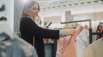 Wall Mural - Beautiful Smiling Female Customer Shopping in Clothing Store, Using Smartphone, Browsing Online, Comparing on Internet, Choosing Stylish Clothes. Fashionable Shop, Colorful Brands, Sustainable Designs