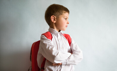 A pensive boy in a school uniform and with a backpack goes to school. Schoolboy indoors on a light background. Free background ready for your lettering. Natural emotions.