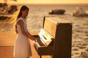 Beautiful girl in a white dress plays the piano on the pier by the sea at sunset