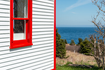 A white narrow wooden clapboard cape cod siding exterior corner wall of a house with bright red trim. There's a closed glass double hung window with red wood trim. A blue sky and ocean in the distance