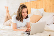 © kalinichenkod - A young businesswoman looks at the screen of a laptop working on the Internet. The girl is lying on the bed and using a laptop