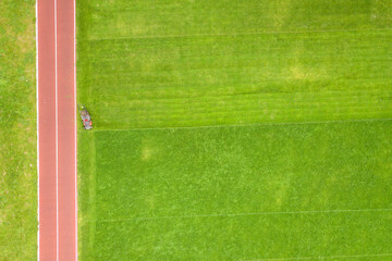 Wall Mural - Aerial view of small figure of worker cutting green grass with mowing mashine on football stadium field with red running tracks in summer.