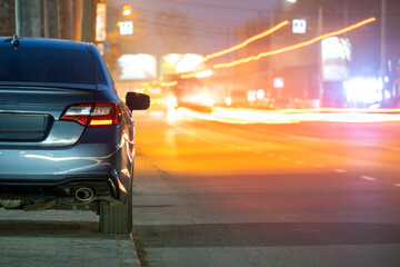 Close up of parked car on roadside at night with blurred view of traffic lights of moving vehicles on city street.