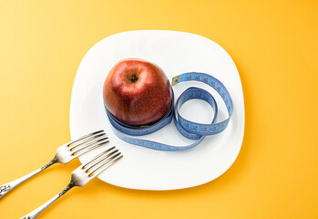 A red apple, wrapped in a centimeter, on a white plate, on a orange background, next to two table forks. The concept of healthy food, fitness, diet, weight loss. View from above. Selective focus.