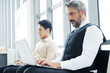 © cocorattanakorn - Serious Senior Caucasian businessman is working with laptop in workspace at office. He sitting distance from asian staff. Technology for business and New normal lifestyle concept.