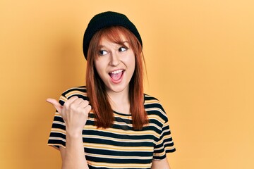 Sticker - Redhead young woman wearing wool cap smiling with happy face looking and pointing to the side with thumb up.