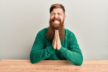 Canvas Print - Young irish redhead man wearing casual clothes sitting on the table praying with hands together asking for forgiveness smiling confident.