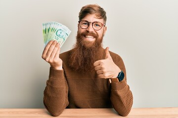 Sticker - Young irish redhead man holding 5 singapore dollars banknotes sitting on the table smiling happy and positive, thumb up doing excellent and approval sign